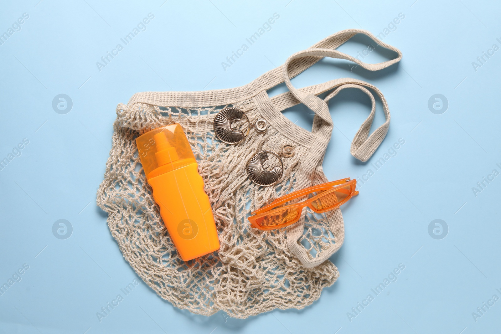 Photo of String bag, sunscreen, sunglasses and earrings on light blue background, top view