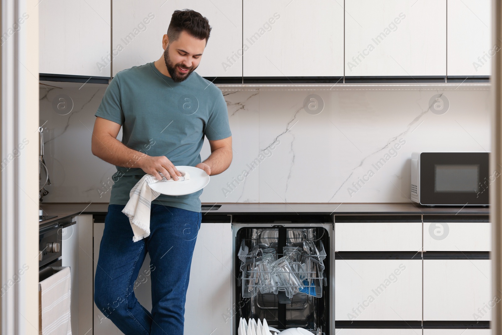 Photo of Smiling man wiping plate near open dishwasher in kitchen