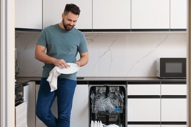 Photo of Smiling man wiping plate near open dishwasher in kitchen