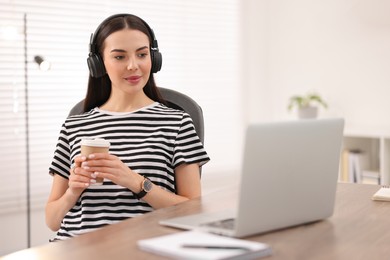 Photo of Young woman in headphones watching webinar at table in room