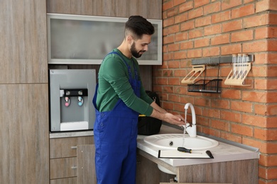 Photo of Male plumber in uniform checking faucet in kitchen. Repair service