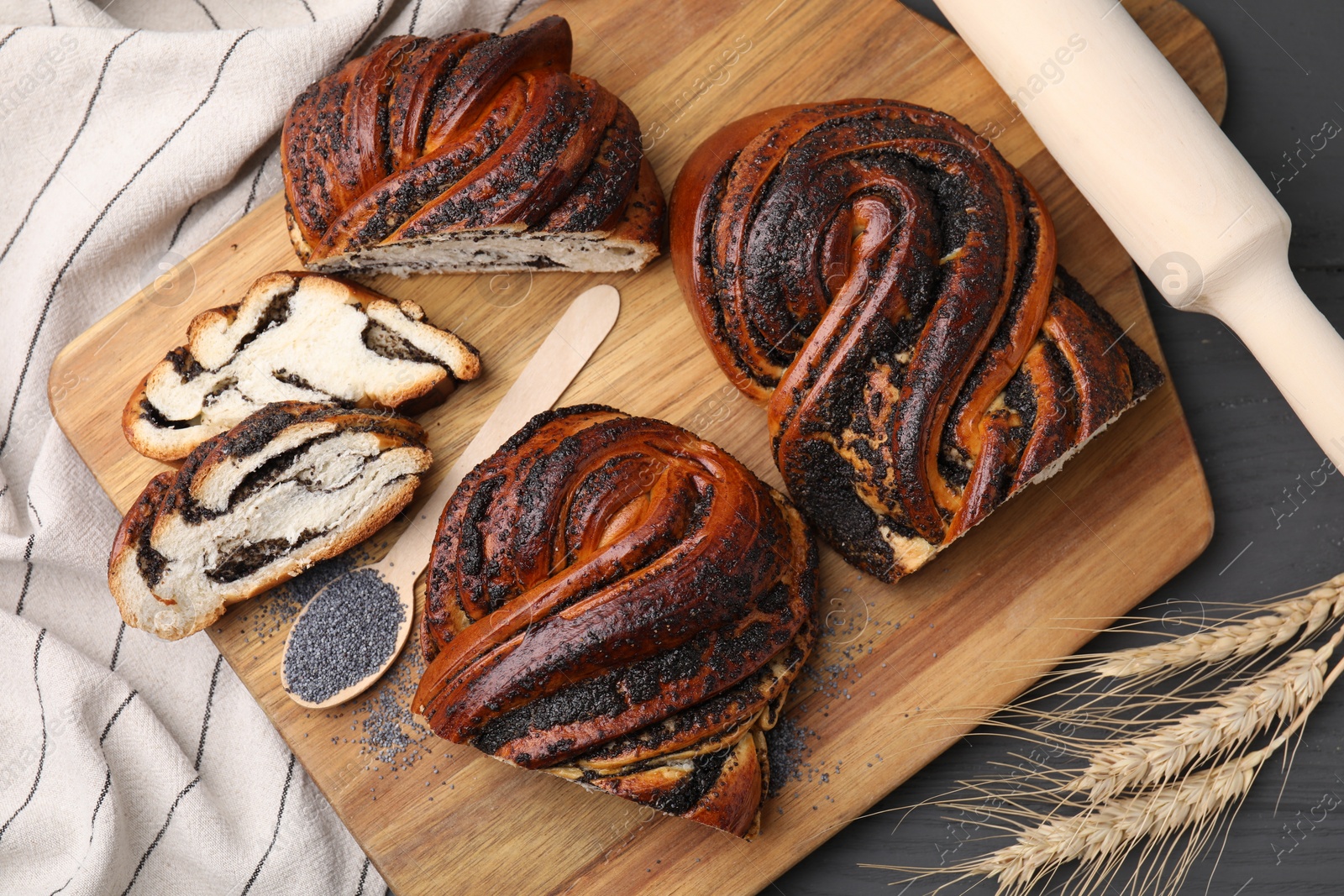 Photo of Pieces of poppy seed roll, spoon and spikelets on grey wooden table, flat lay. Tasty cake