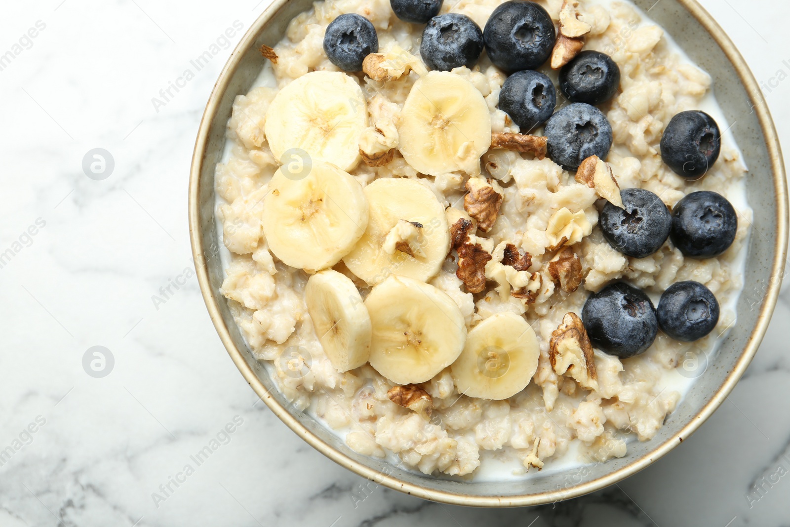 Photo of Tasty oatmeal with banana, blueberries, walnuts and milk served in bowl on white marble table, top view