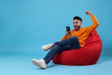 Photo of Happy young man using smartphone on bean bag chair against light blue background. Space for text