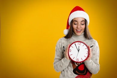 Woman in Santa hat with clock on yellow background, space for text. New Year countdown