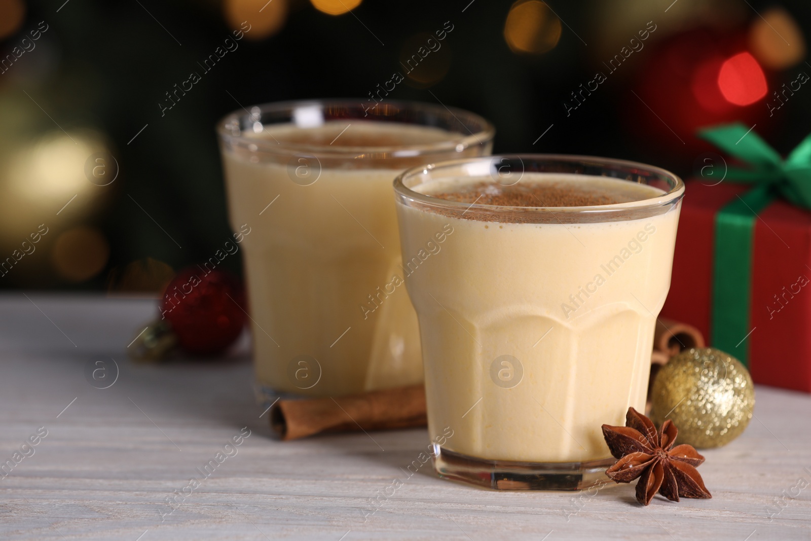 Photo of Tasty eggnog, cinnamon, anise and Christmas decorations on white wooden table against blurred lights