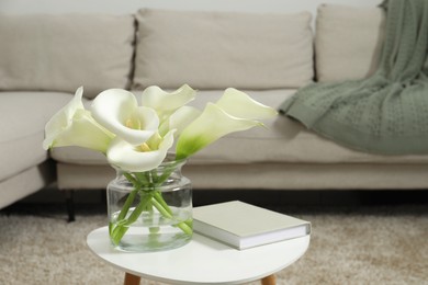 Photo of Beautiful calla lily flowers in glass vase and book on white table indoors