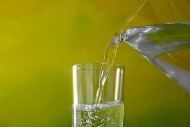 Photo of Pouring water from jug into glass on color background, closeup