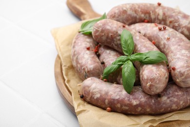 Board with raw homemade sausages, basil leaves and peppercorns on white tiled table, closeup. Space for text