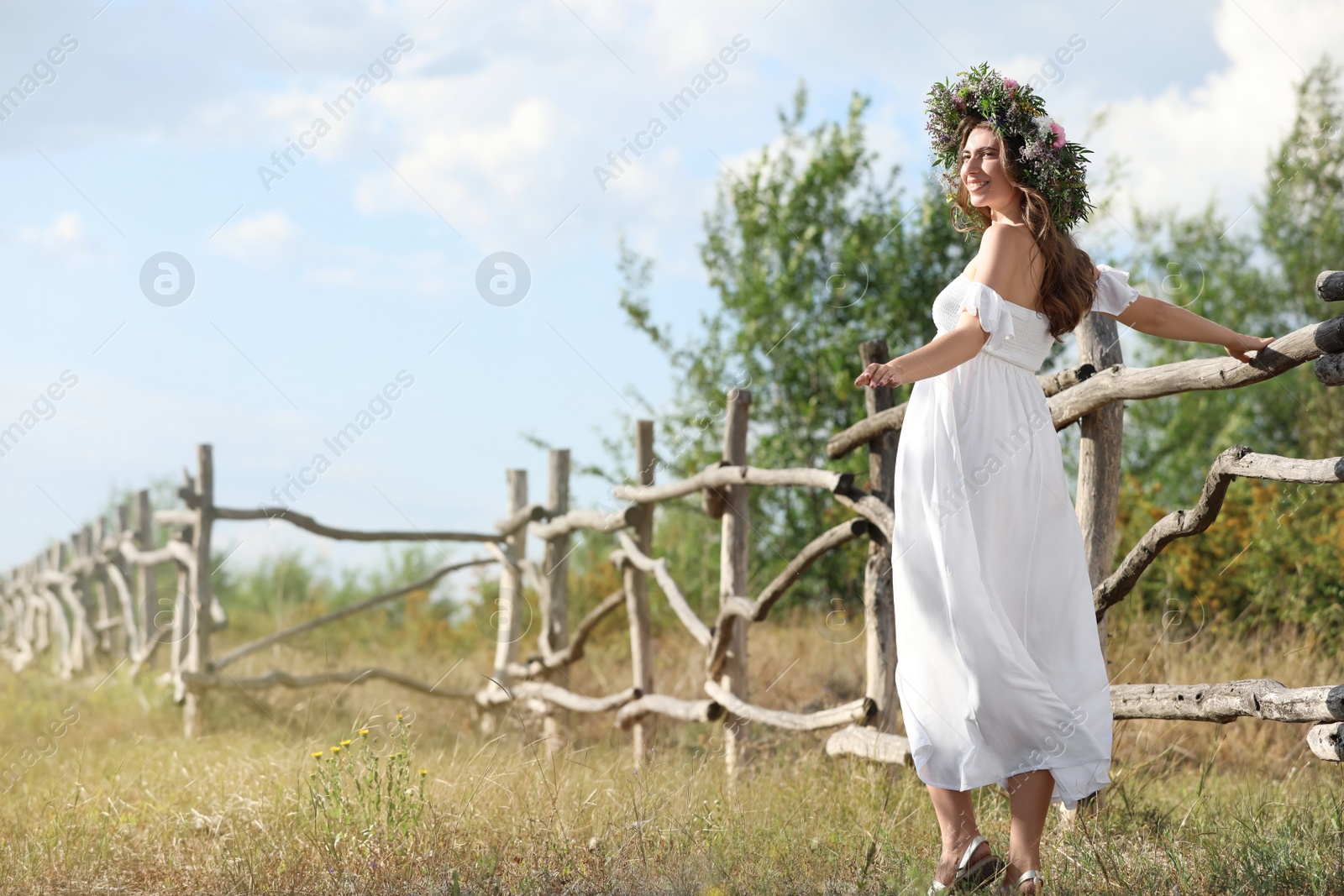 Photo of Young woman wearing wreath made of beautiful flowers near wooden fence on sunny day