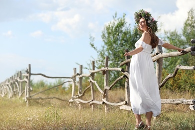 Young woman wearing wreath made of beautiful flowers near wooden fence on sunny day