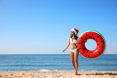 Young woman wearing Santa hat and bikini with inflatable ring on beach, space for text. Christmas vacation