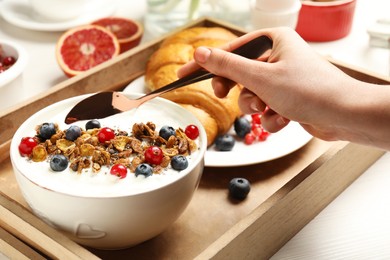 Photo of Woman eating yogurt with granola and berries at white wooden table, closeup