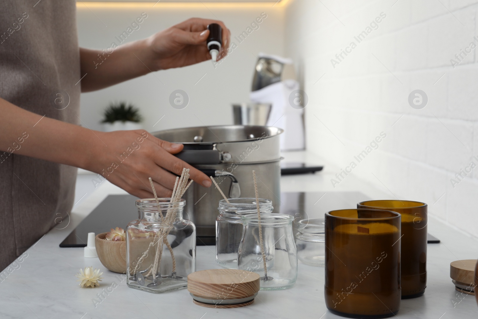 Photo of Woman adding essential oil into pot with melted wax on stove in kitchen, closeup. Making homemade candles