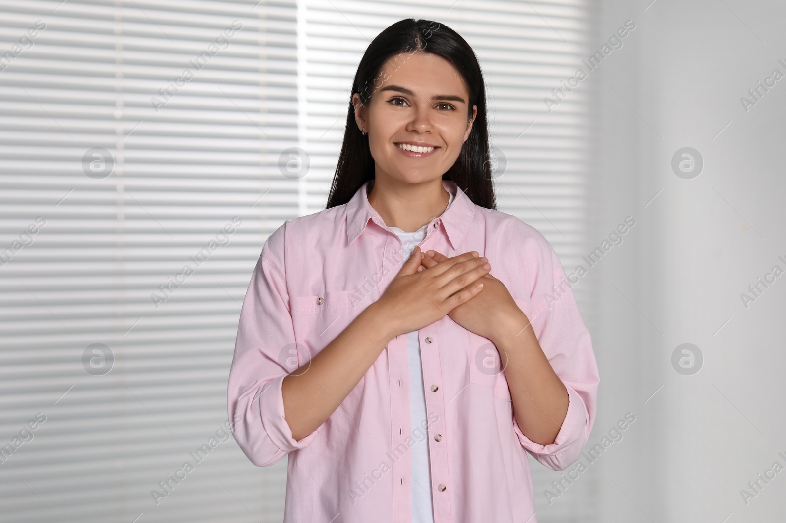 Photo of Thank you gesture. Beautiful grateful woman holding hands near heart indoors