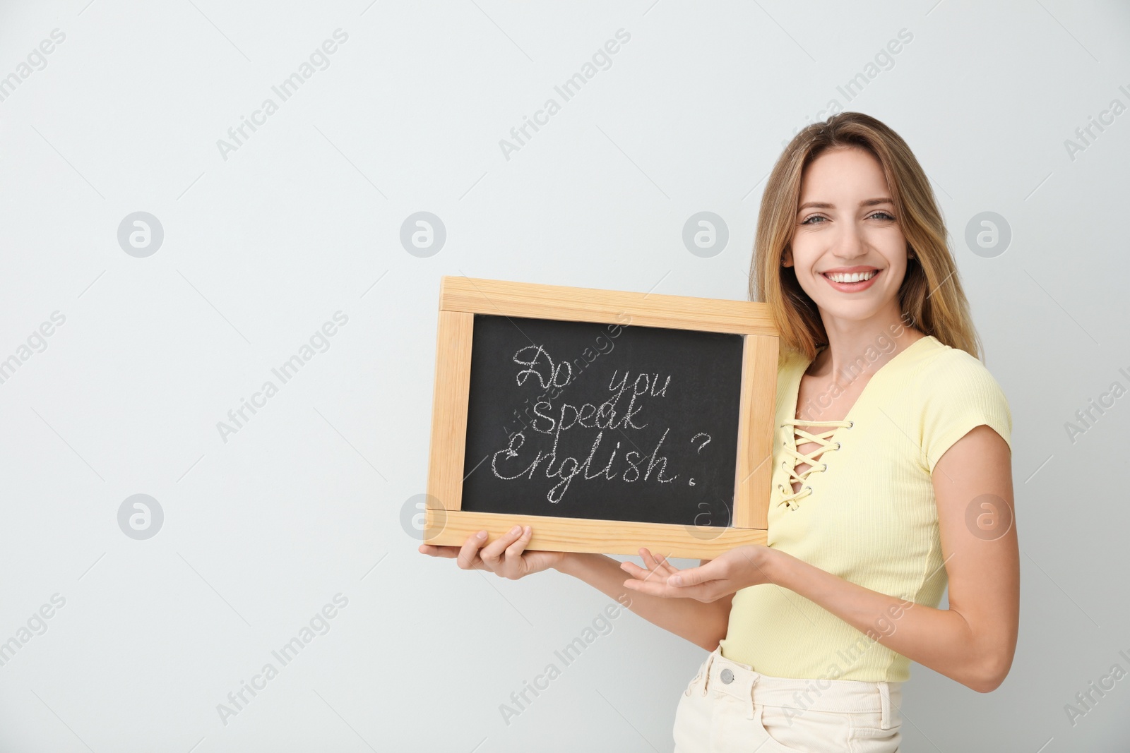 Photo of Young female teacher holding chalkboard with words DO YOU SPEAK ENGLISH? on light background. Space for text