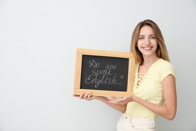 Photo of Young female teacher holding chalkboard with words DO YOU SPEAK ENGLISH? on light background. Space for text