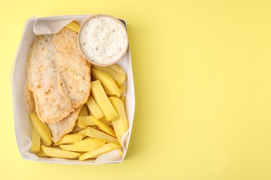 Delicious fish and chips with tasty sauce in paper box on yellow table, top view. Space for text