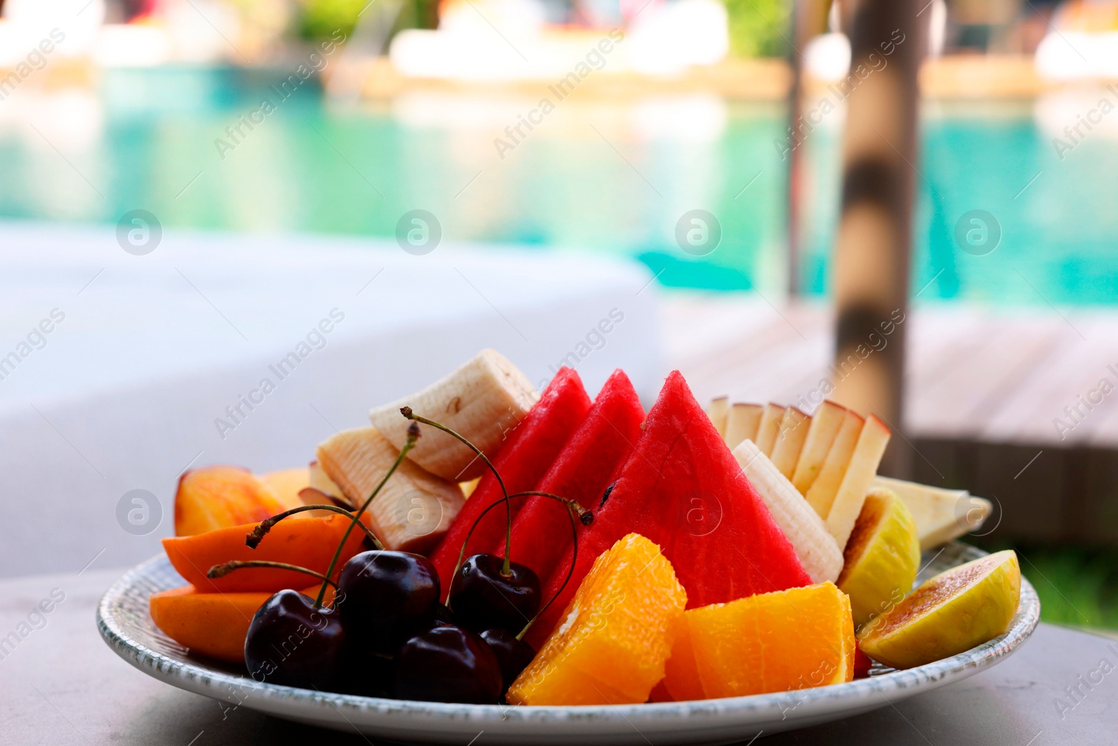 Photo of Plate with fresh fruits on table near sun lounger. Luxury resort with outdoor swimming pool