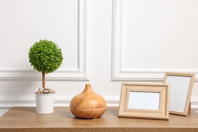 Green artificial plant in pot, frames and air humidifier on wooden table near white wall