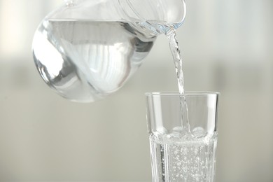 Pouring water from jug into glass on blurred background, closeup