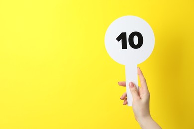 Photo of Woman holding auction paddle with number 10 on yellow background, closeup. Space for text