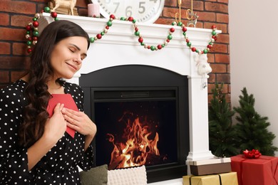 Young woman with greeting card sitting near fireplace indoors