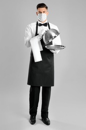 Photo of Waiter in medical face mask with empty tray on light grey background