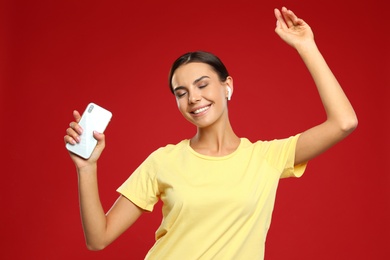 Happy young woman with smartphone listening to music through wireless earphones on red background