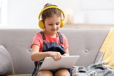 Photo of Cute little girl with headphones and tablet on sofa at home