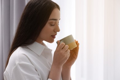 Photo of Beautiful woman with cup of drink near window at home, space for text. Lazy morning