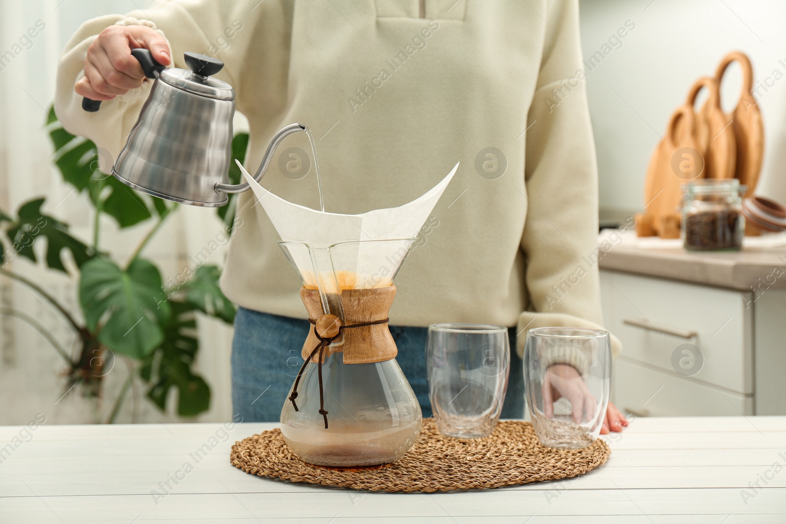 Photo of Woman pouring hot water into glass chemex coffeemaker with paper filter and coffee at white table, closeup