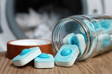 Jar with water softener tablets on wooden table near washing machine, closeup