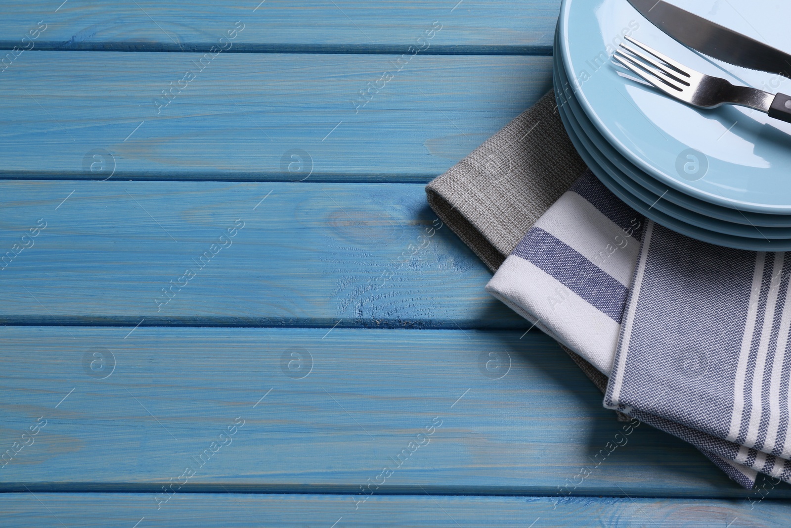 Photo of Different kitchen towels and stack of plates with cutlery on blue wooden table. Space for text