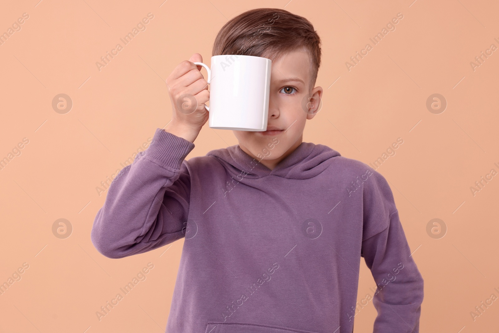 Photo of Cute boy covering eye with white ceramic mug on beige background