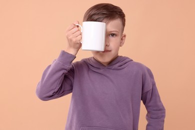 Photo of Cute boy covering eye with white ceramic mug on beige background