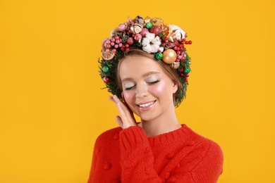 Beautiful young woman wearing Christmas wreath on yellow background