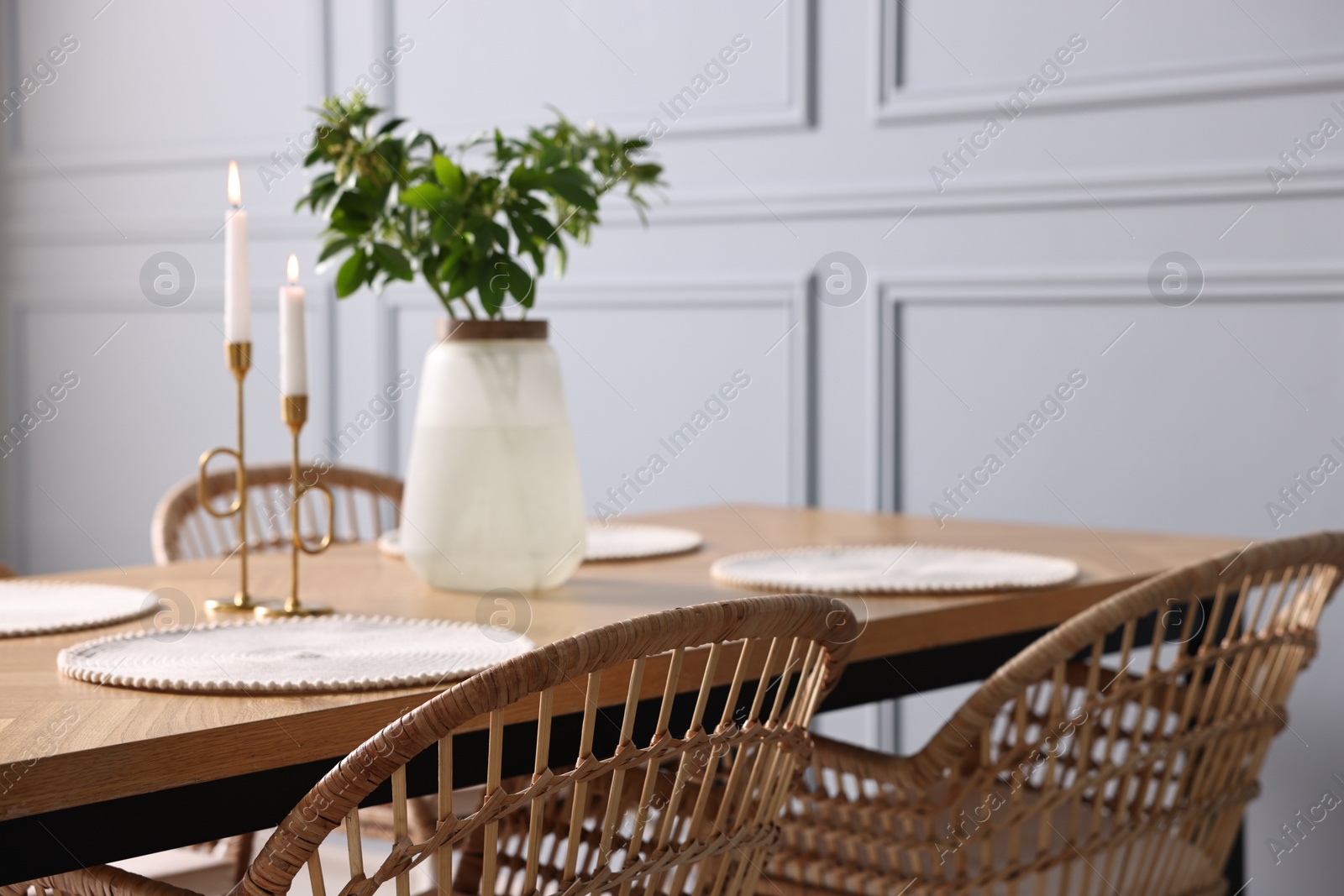 Photo of Rattan chairs, table, burning candles and vase with green branches in stylish dining room