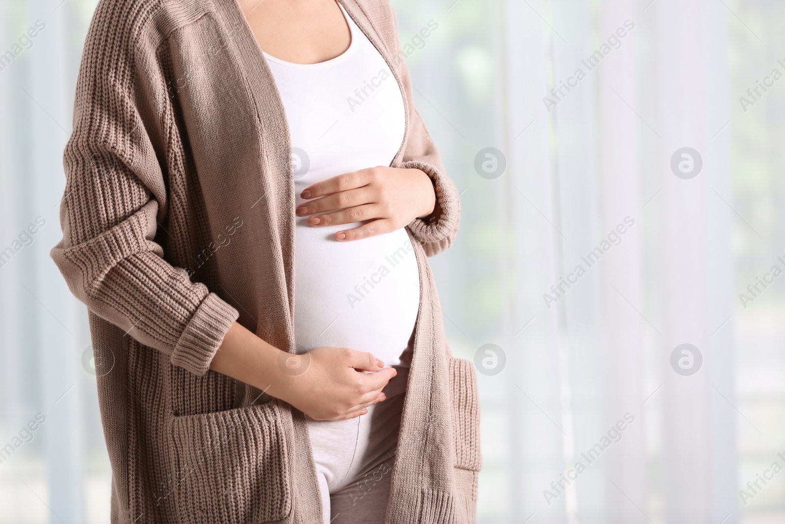 Photo of Pregnant woman standing near window at home, closeup