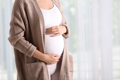 Pregnant woman standing near window at home, closeup