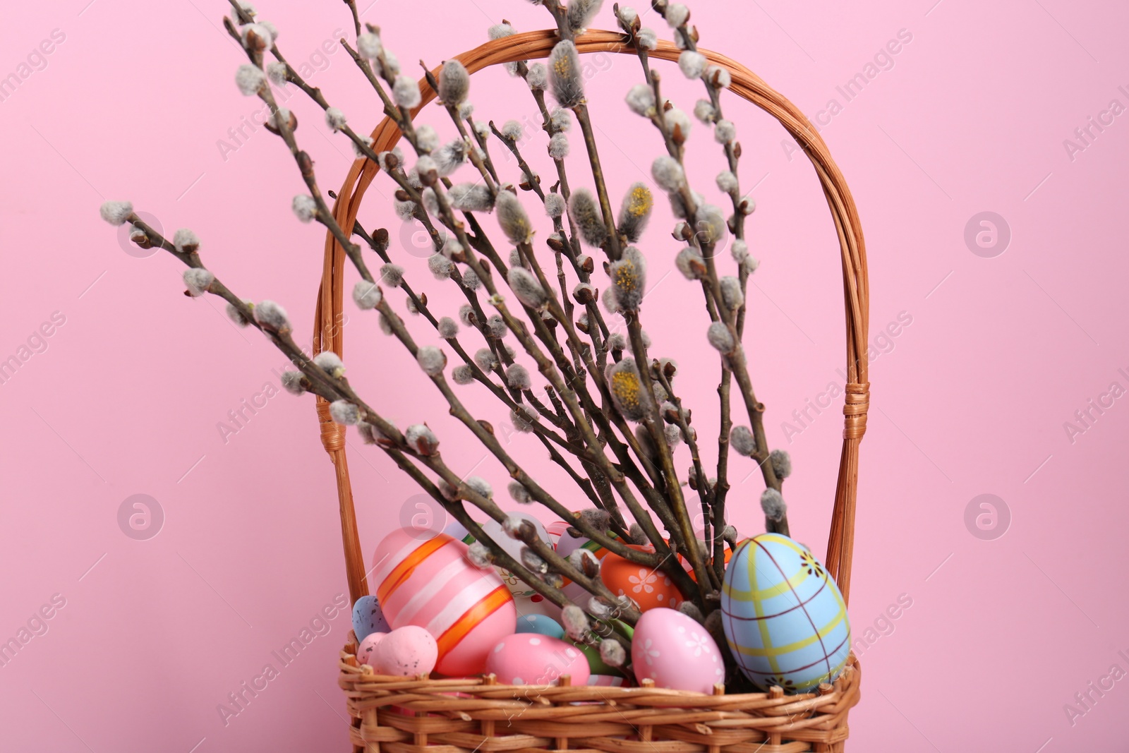 Photo of Wicker basket with beautiful willow branches and painted eggs on pink background. Easter decor