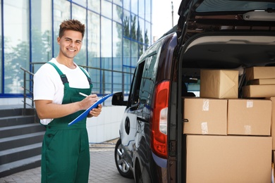 Photo of Young courier holding clipboard near delivery van with parcels outdoors