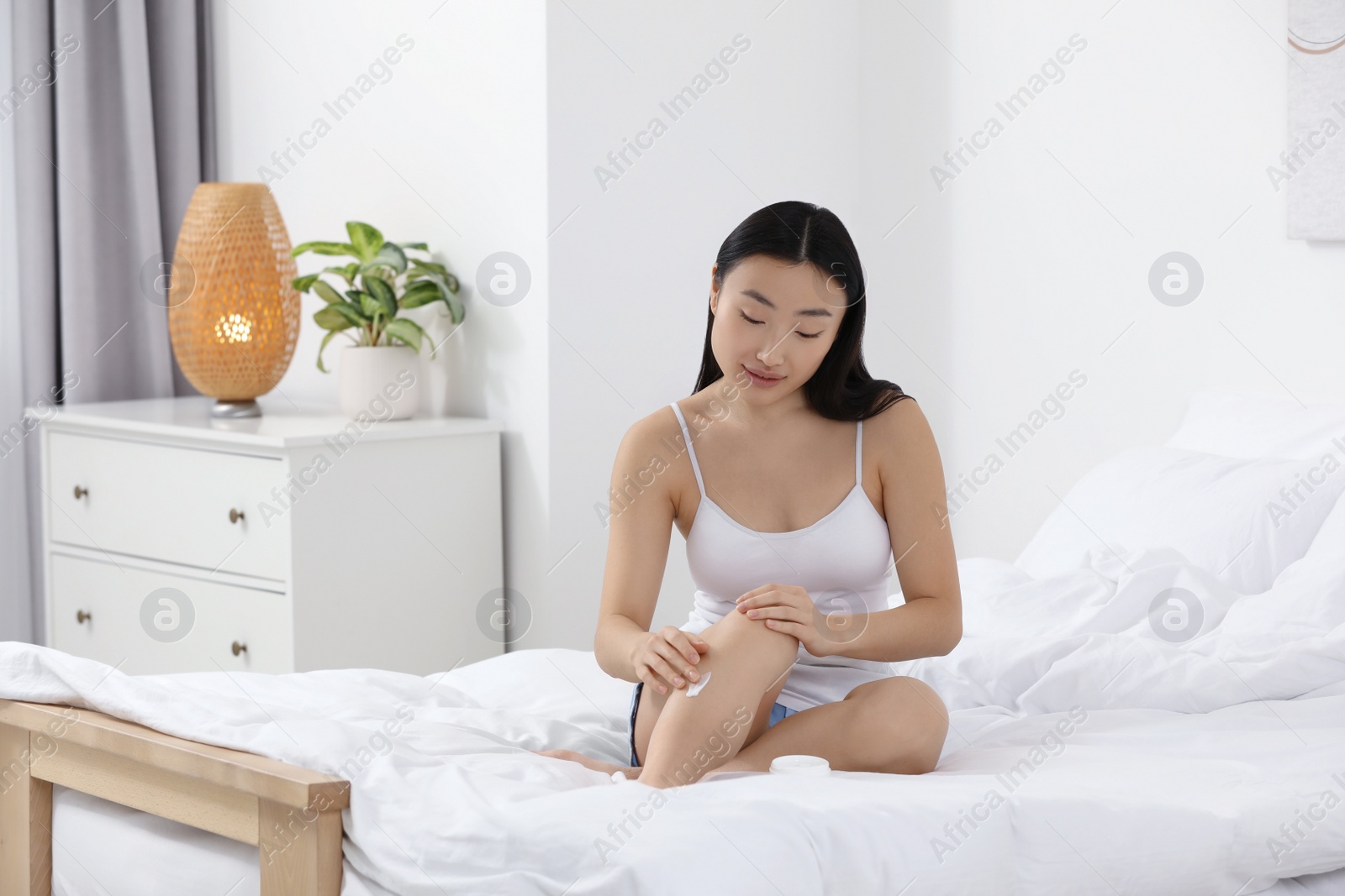 Photo of Beautiful young Asian woman applying body cream on leg in bedroom