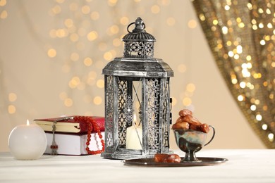 Photo of Arabic lantern, Quran, misbaha, candle and dates on white table against blurred lights