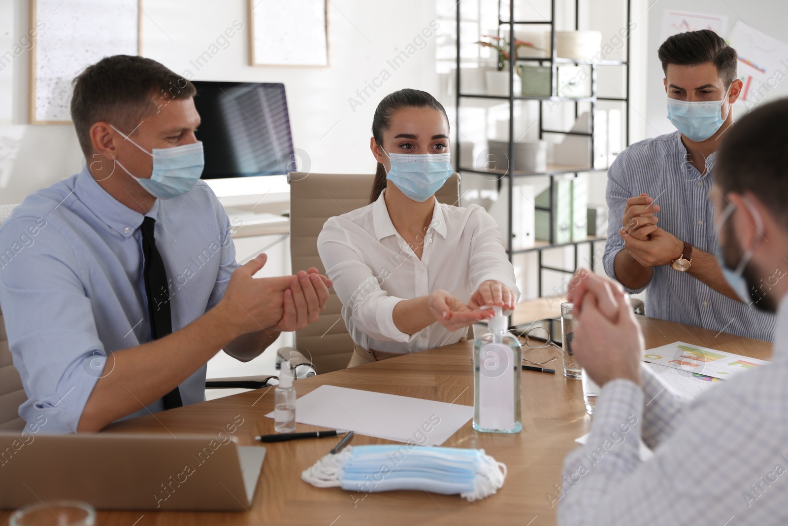 Photo of Coworkers with protective masks using hand sanitizer in office. Business meeting during COVID-19 pandemic