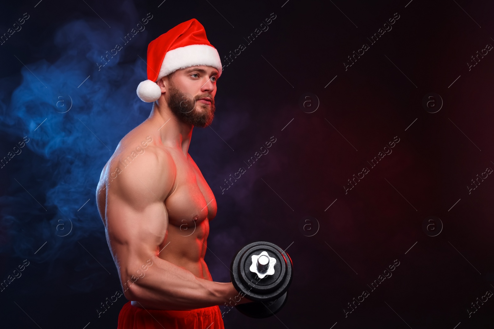 Photo of Muscular young man in Santa hat with dumbbell on black background, space for text