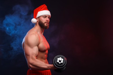 Photo of Muscular young man in Santa hat with dumbbell on black background, space for text