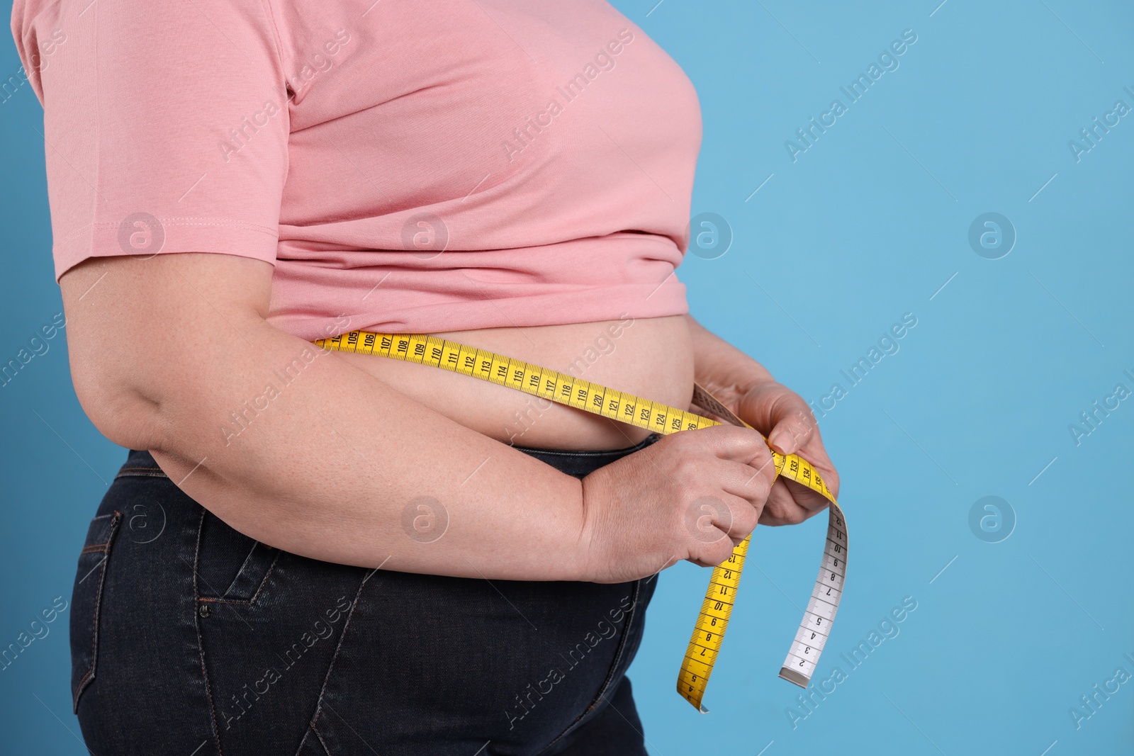 Photo of Overweight woman measuring waist with tape on light blue background, closeup