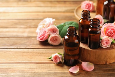 Photo of Bottles of rose essential oil and fresh flowers on wooden table, space for text