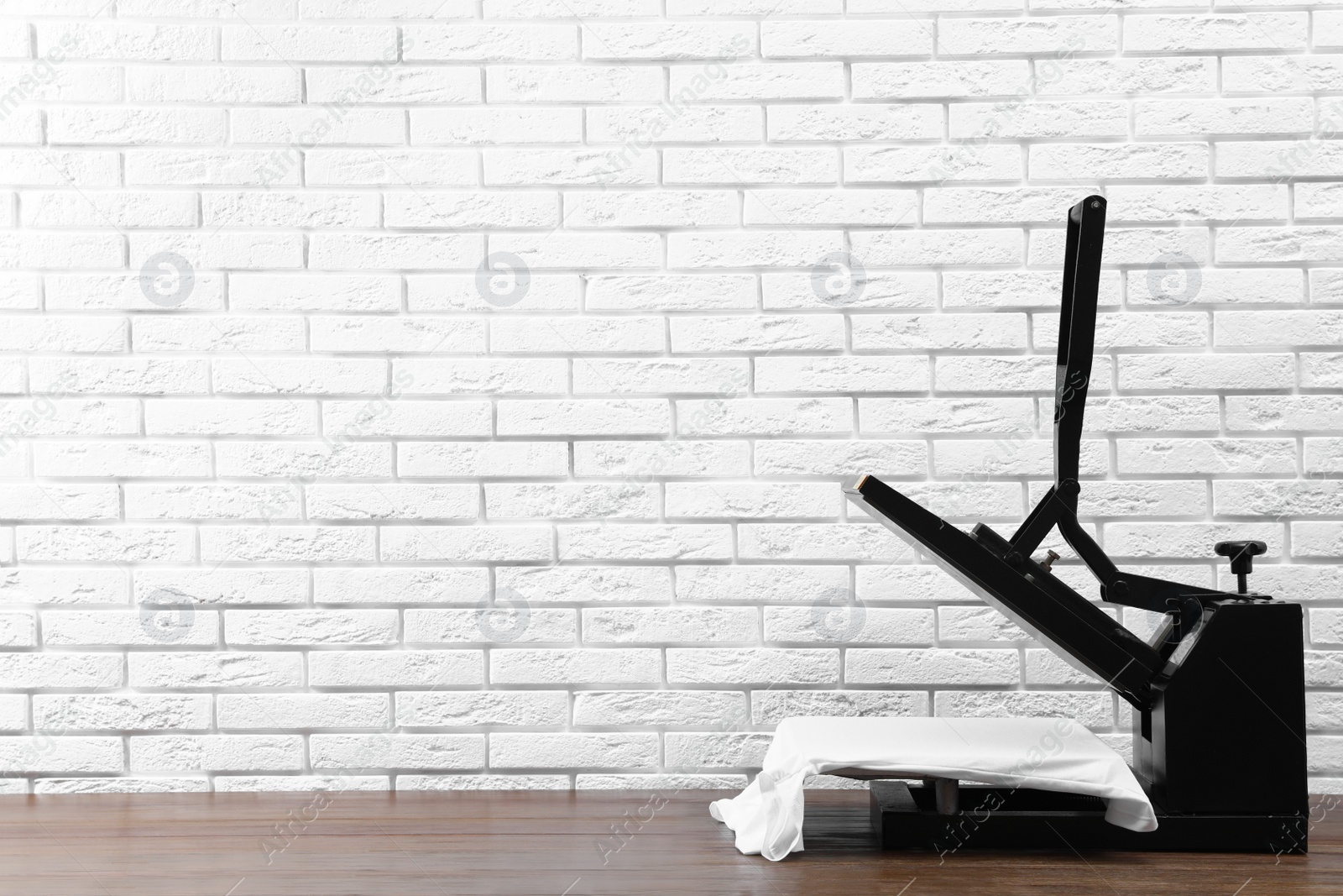 Photo of Heat press machine with t-shirt on wooden table near white brick wall, space for text